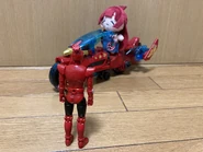 Peluche de Yaku con una moto observada por un muñeco.