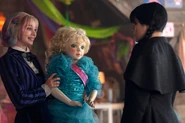 Enid Sinclair holding a doll with blonde hair and a blue dress with Wednesday Addams standing in front of her