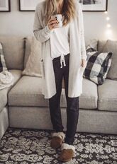 Photo of a woman in a neutral-colored living room. She is wearing a gray cashmere sweater coat with a white t-shirt, black sweatpants, and brown slippers with fluff on the inside. She is also holding a mug with a letter on it.