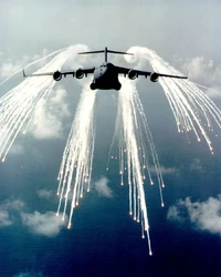 A C-17/91 releases a barrage of flares. The wing-tip vortices can be seen in the flare smoke behind the aircraft.