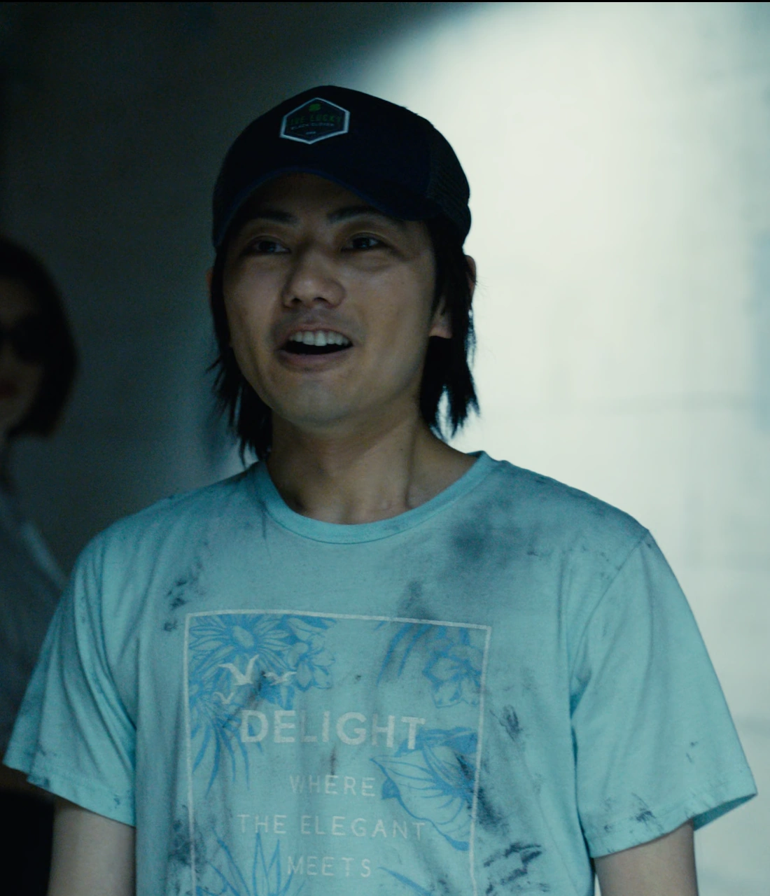 A screenshot of Tatta Kodai from Season 1, a young man with long hair, wearing a blue T-shirt and a cap, happily looking at someone in front of him