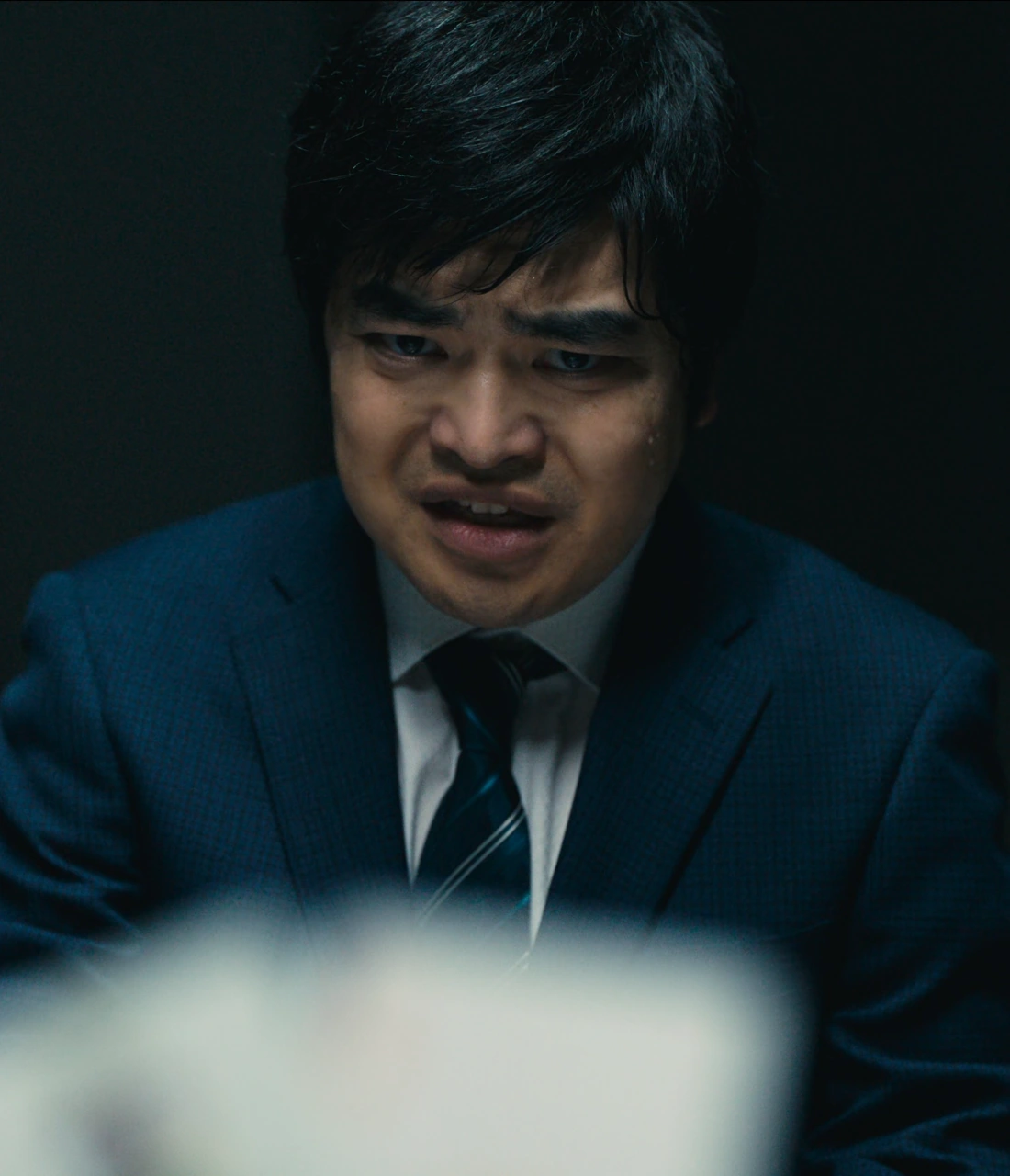 A screenshot of an unnamed player from Season 3, a man with short black hair, wearing a classic blue suit with a tie, looking at his opponent with panic