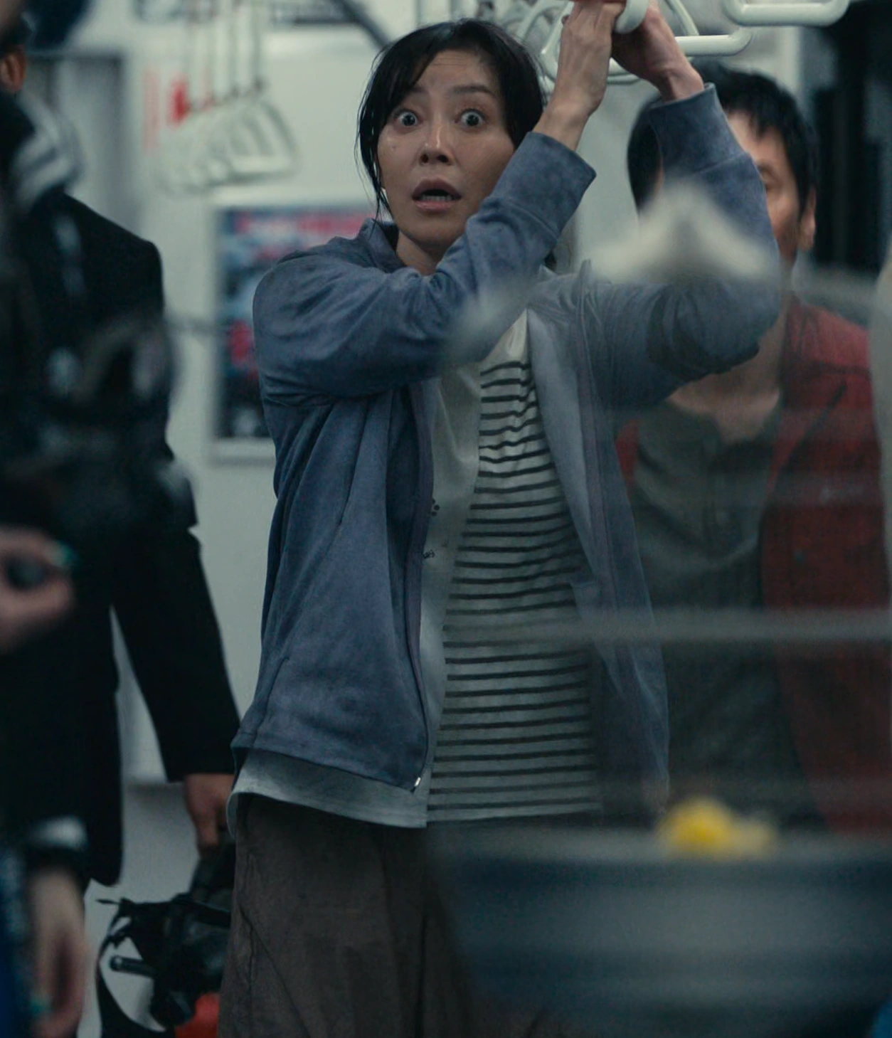 A screenshot of Sachiko Makino from Season 3, an older woman in a striped shirt and light jacket, gripping a handrail with a startled expression