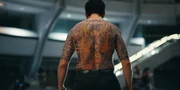 Kazuya stands shirtless with his back to the camera, revealing an elaborate full-back tattoo of a fierce dragon in red, yellow, and black ink