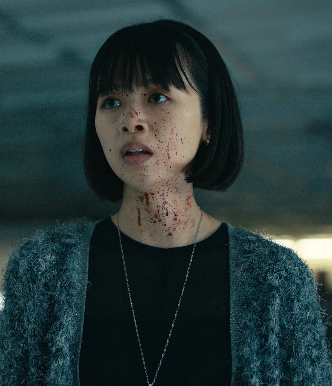 A middle-aged woman, named Asami in the subtitles, with bob-style hair and bangs, dressed in a black shirt with a woolen green cardigan on top and a gray skirt