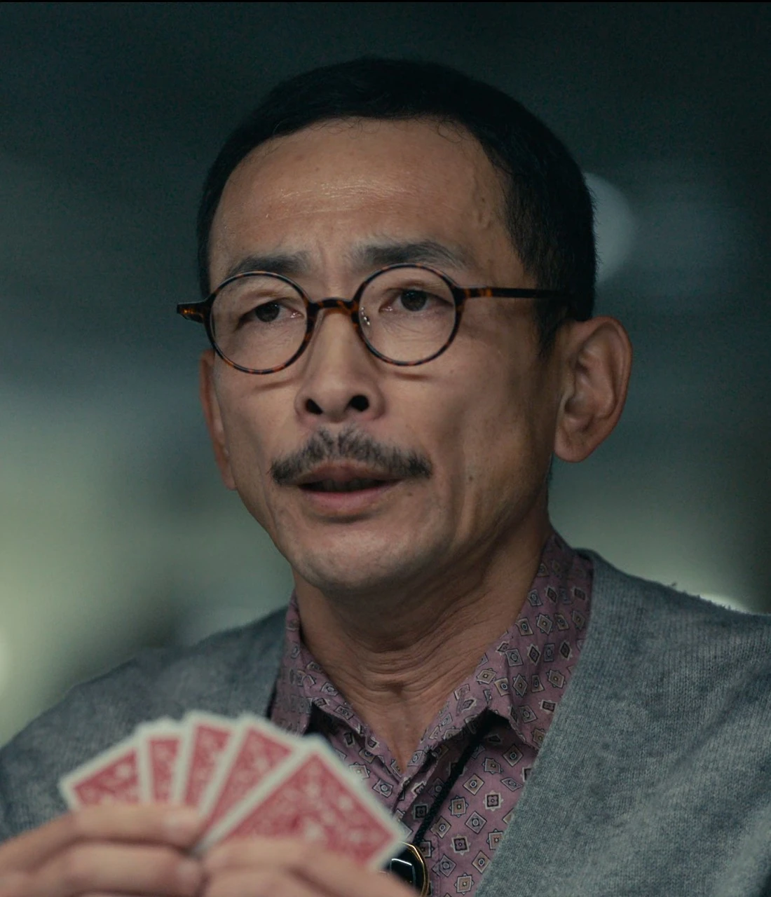 An elderly man with a moustache in round glasses, wearing a pink shirt with a gray cardigan on top