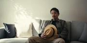 Aguni sitting on a sofa, holding Danma's hat in his hands
