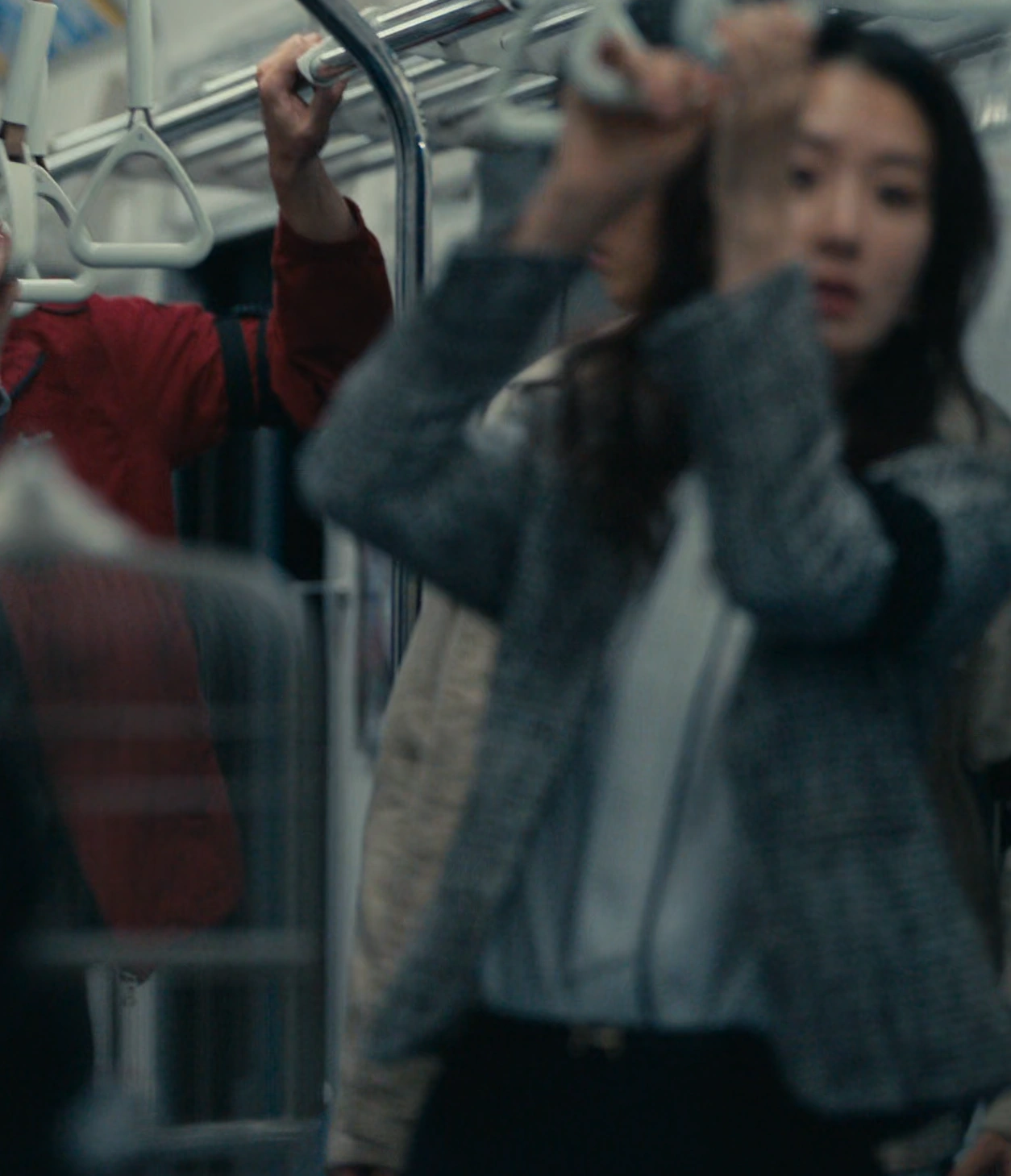 A screenshot of Shion from Season 3, a young woman with shoulder-length straight hair in a gray coat, holding onto a strap while looking ahead with concern