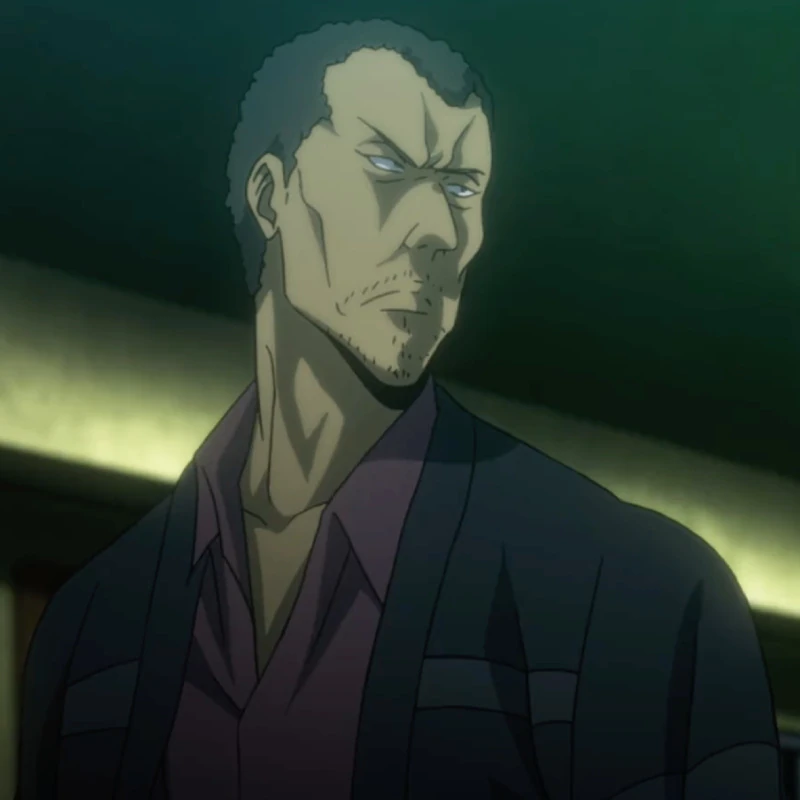 A dark-skinned man with a short afro, a stubbly beard, and dressed in a dull purple collar shirt with a black open cardigan on top