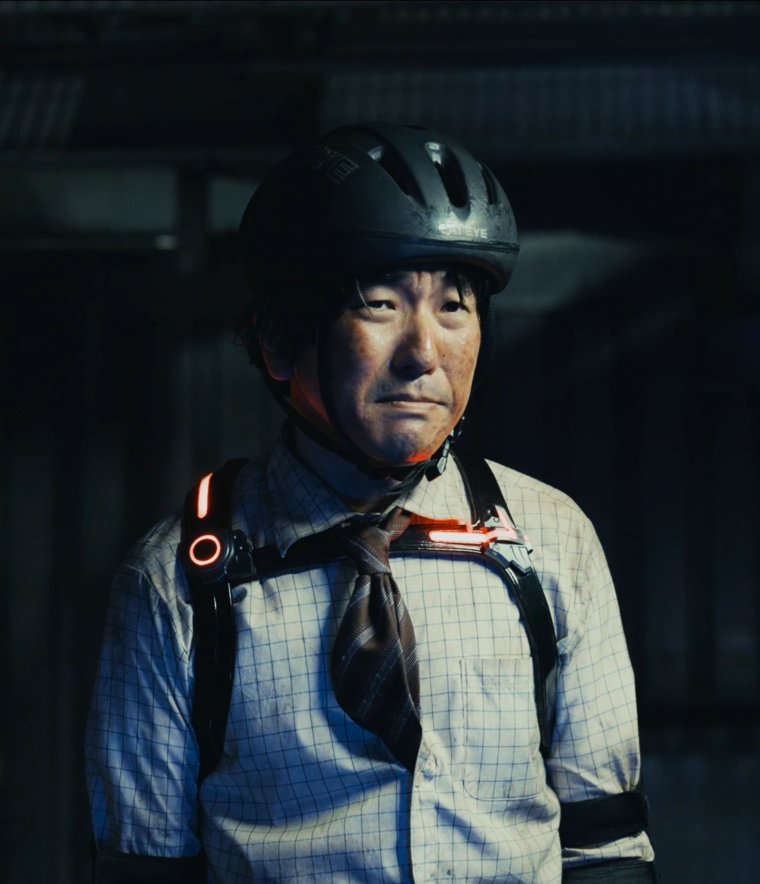 A screenshot of Fujiki from Season 2, a man wearing a bike helmet, a white grid-patterned shirt, and a brown striped tie