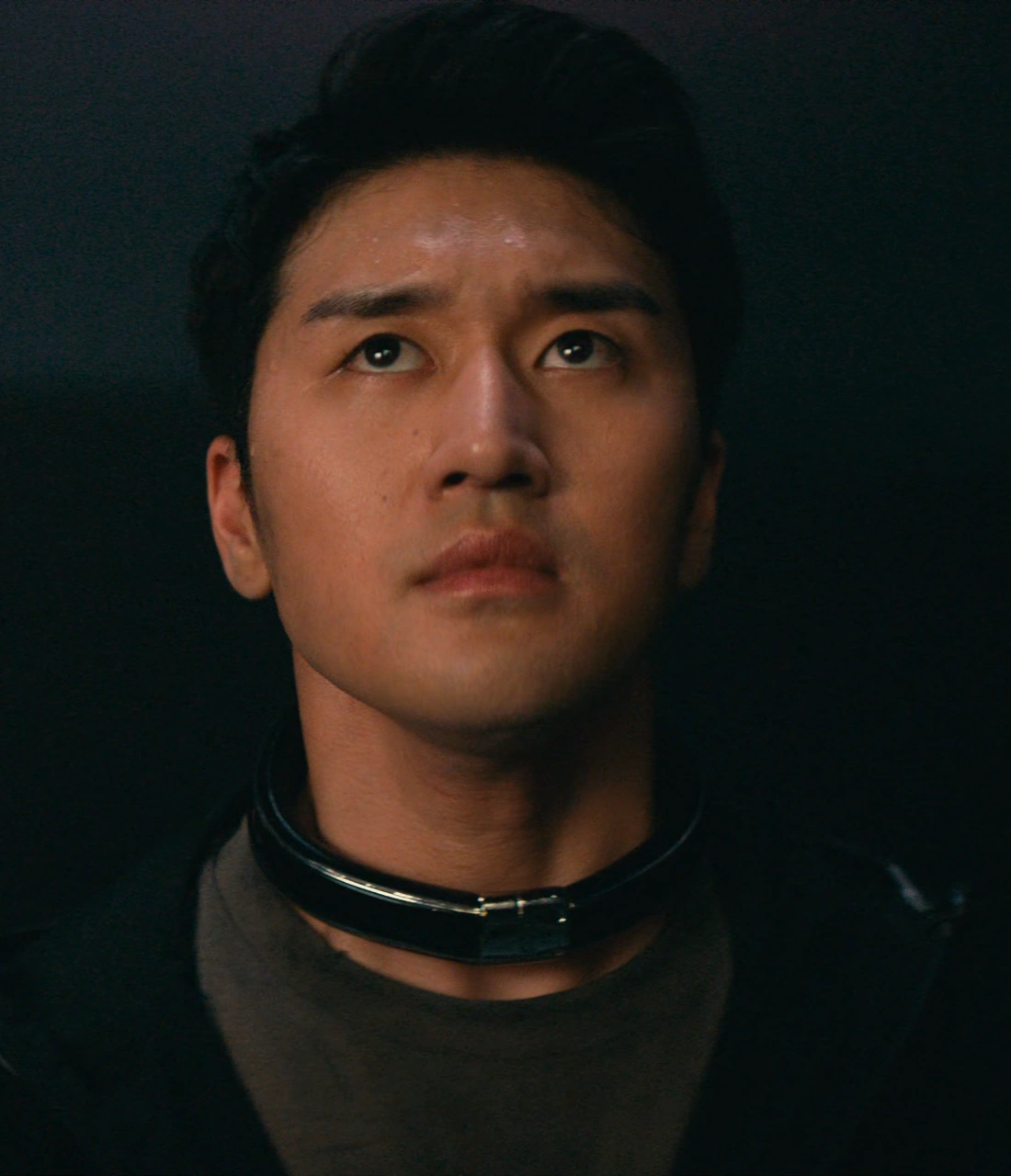 A close-up shot of Itsuki gazing upwards with a serious expression, wearing a metal collar around his neck