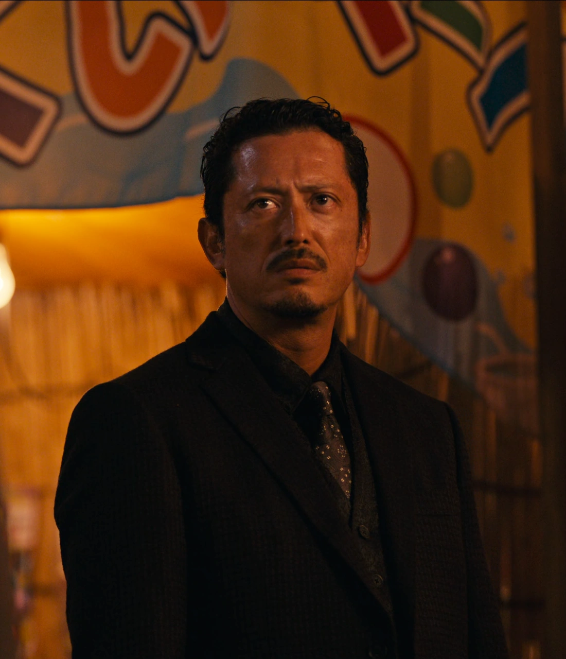 A close-up shot of Kazuya dressed in a black suit and looking around with suspicion
