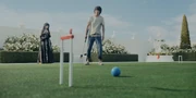 A croquet field with wickets and balls on the grass, Mira standing aside and smiling while Arisu is annoyed after missing the hit