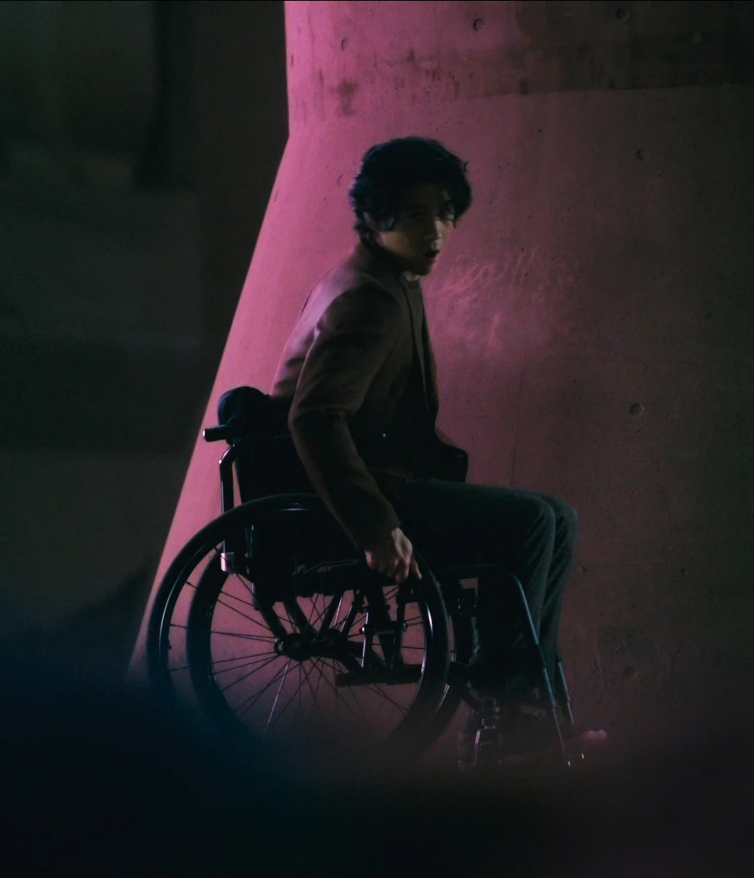 Ryuji in his wheelchair hiding behind a large pillar, illuminated with pink, and talking