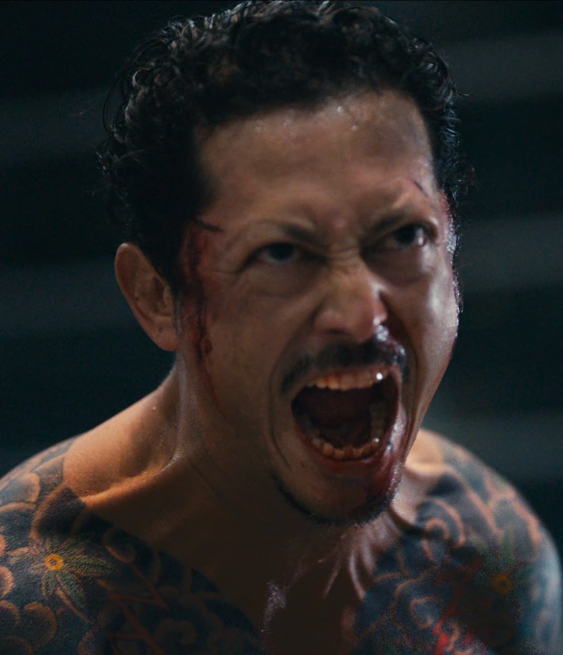 A close-up shot of Kazuya with his bare chest covered in tattoos and blood streaming down his face, he is screaming with rage and determination