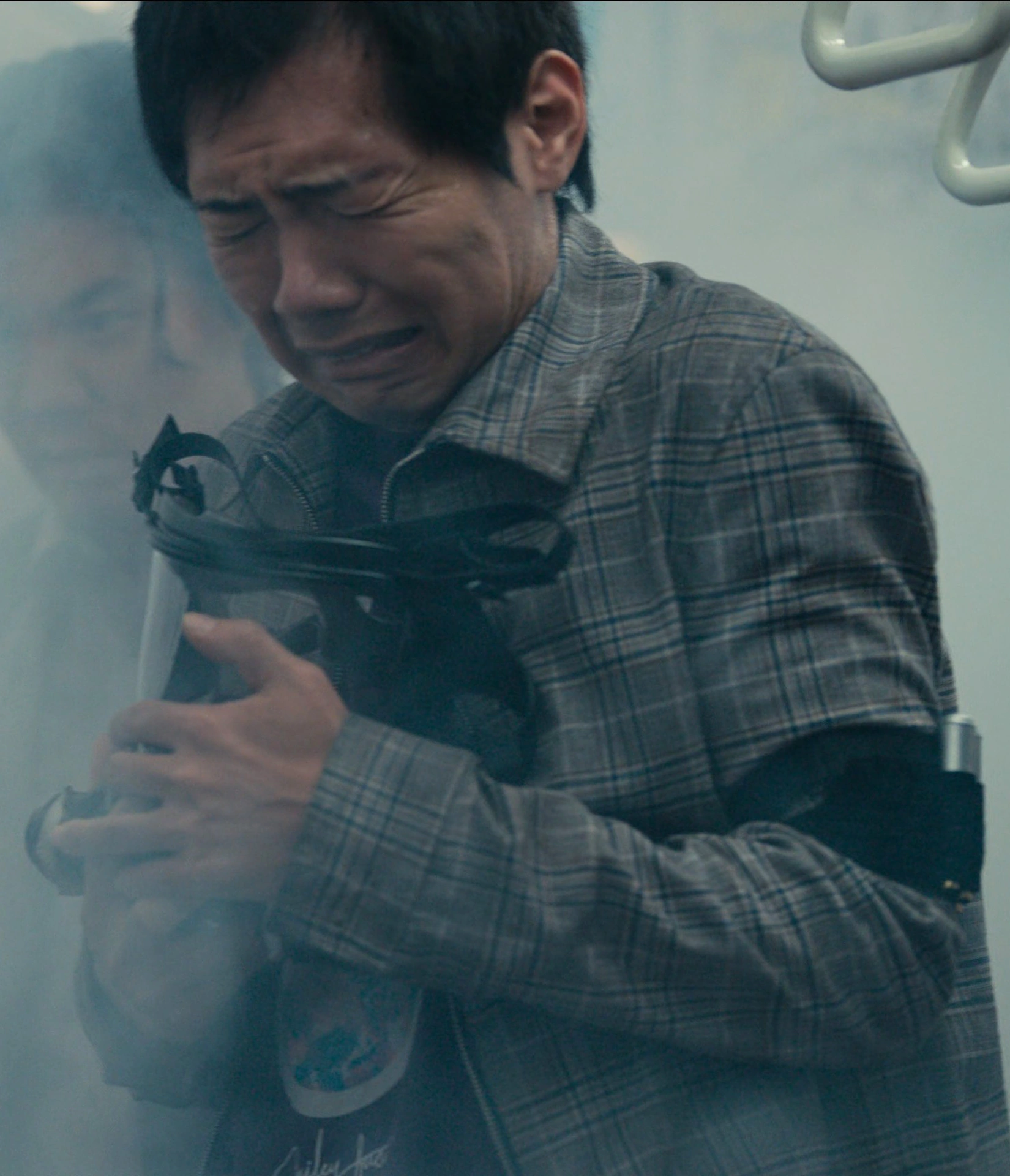 A screenshot of an unnamed male player in a checkered shirt over a purple T-shirt, holding onto his gas mask and crying, as the air around him is filled with unknown substance