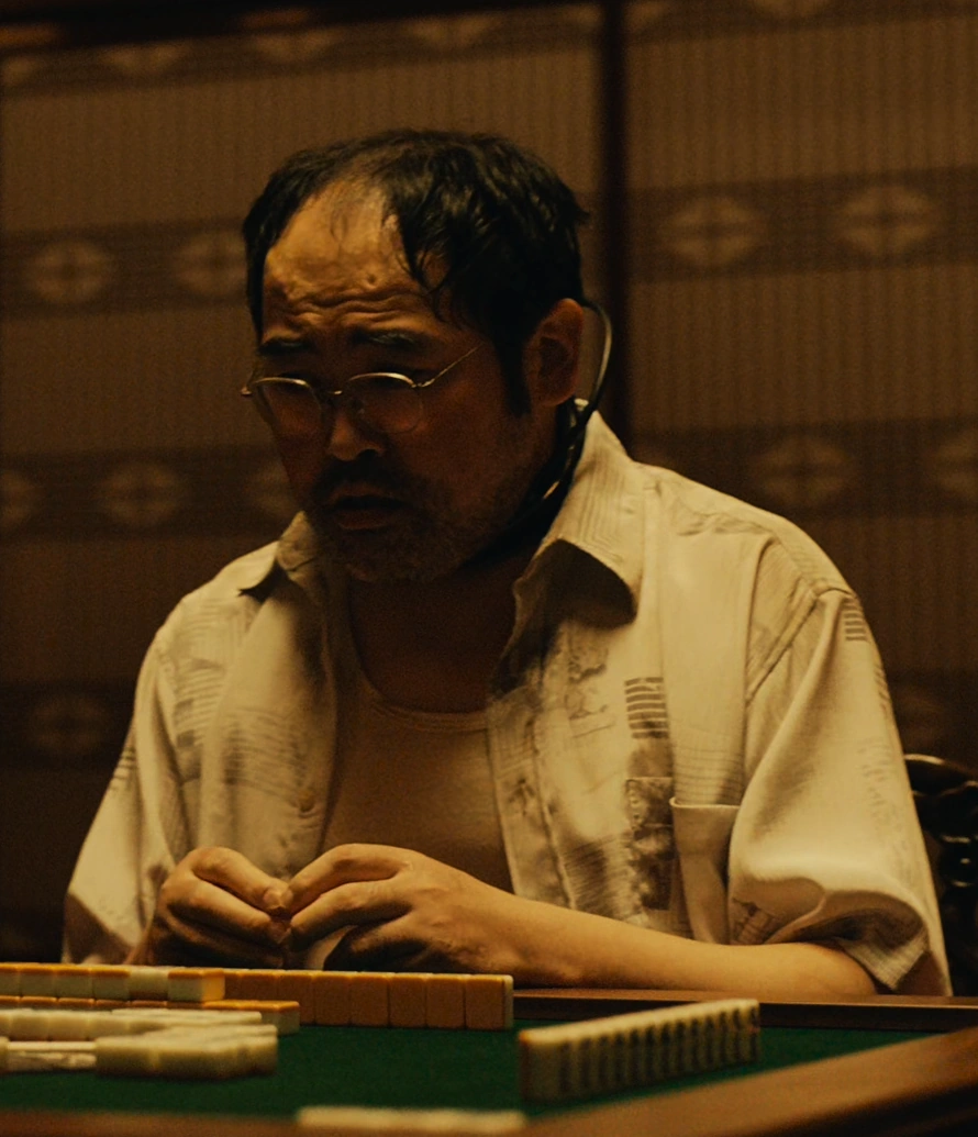A screenshot of an unnamed character from Season 2, an older man in glasses with short receding dark hair, who is wearing a light-colored beach-style shirt over a white T-shirt
