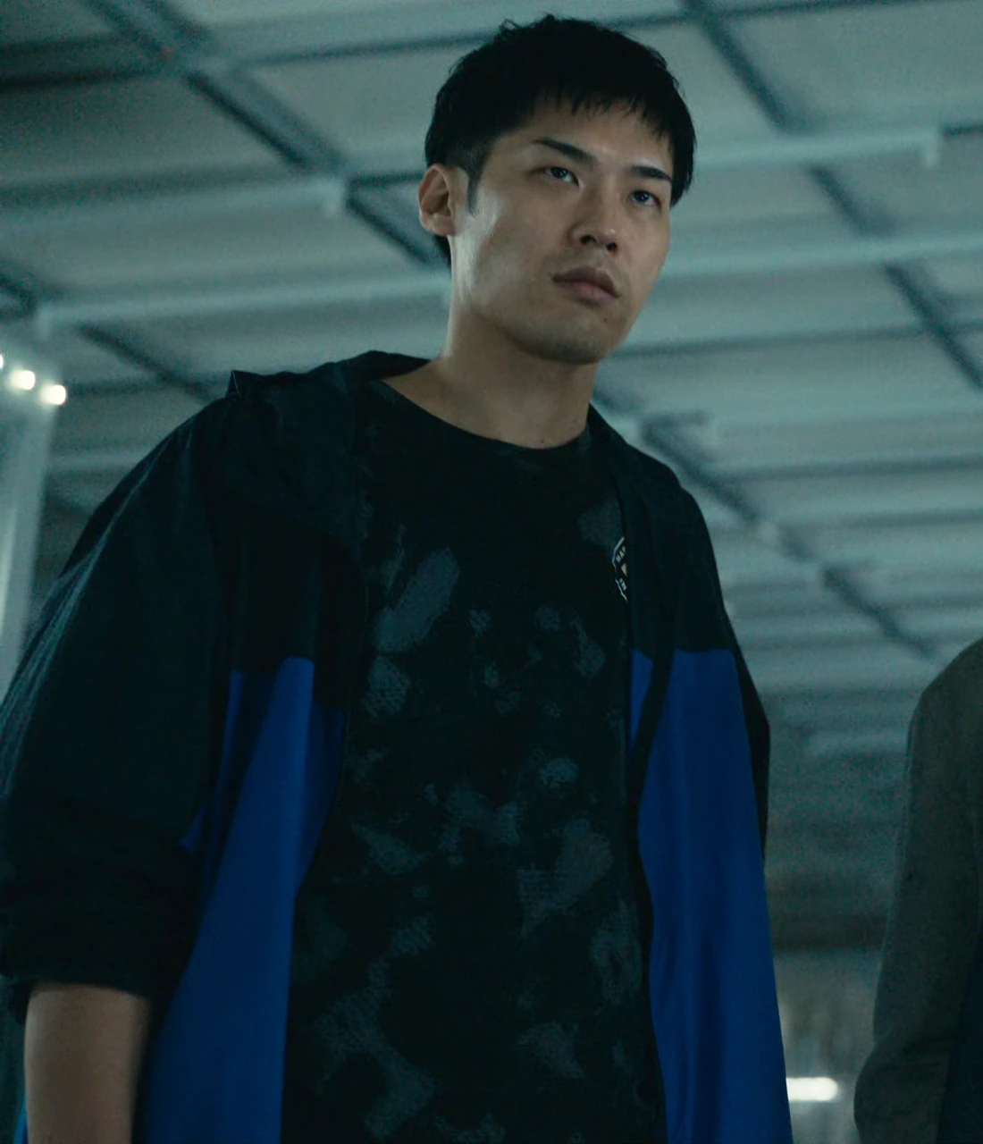 A tall middle-aged man, named Tominaga in the subtitles, wearing a blue and black tracksuit with a dark T-shirt underneath