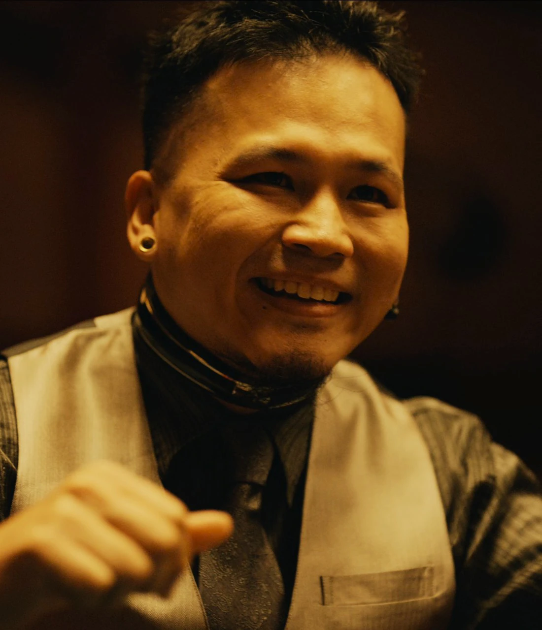 A screenshot of an unnamed character from Season 2, a laughing young man with short hair and a trimmed goatee, who is wearing a dark dress shirt paired with a shiny grey vest and a patterned tie. He has noticeable ear piercings (gauged lobes)