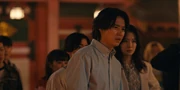 Arisu with a confused and thoughtful expression, standing in the crowd of people with lanterns and a big red gate behind him