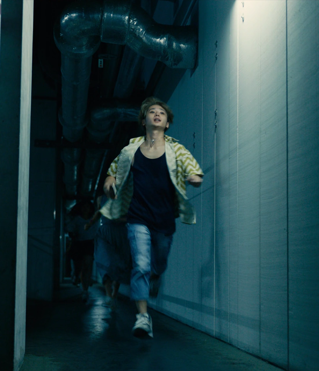Shota, a young man with blond hair, wearing jeans and a yellow shirt on top of a black T-shirt. He is running through a dim corridor while holding his phone