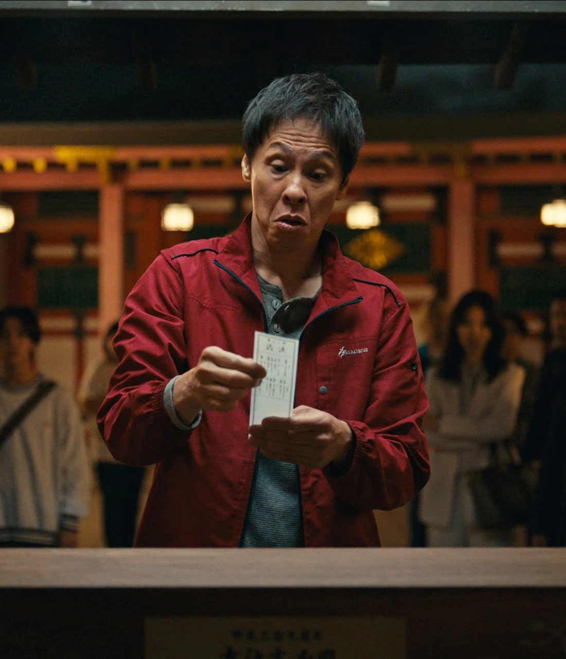 A close-up shot of Tetsu reading his fortune slip with a surprised but pleased expression, multiple other characters are waiting behind him