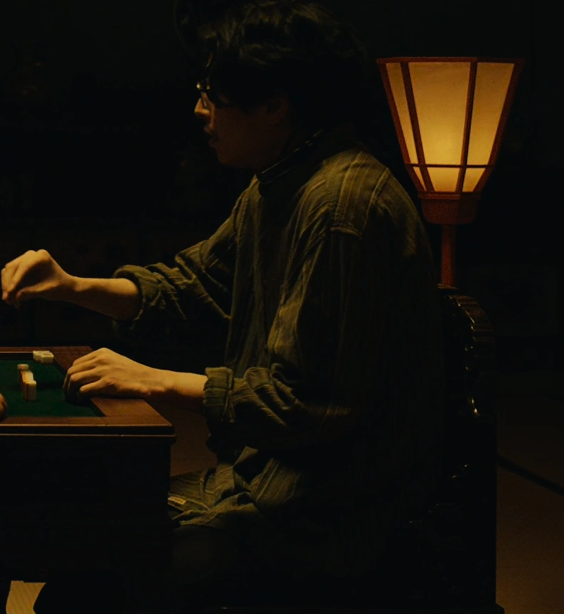 A screenshot of an unnamed character from Season 2, a middle-aged man in glasses with shoulder-length wavy dark hair, wearing a dark shirt and facing the camera with his left side