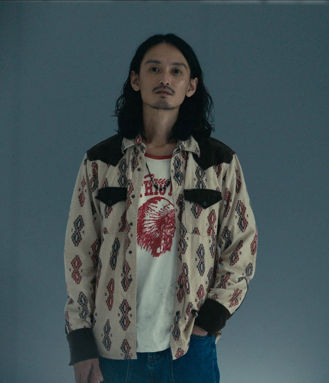 A man with long curly hair, wearing a beige shirt with geometric patterns over a white T-shirt with a red print and blue jeans