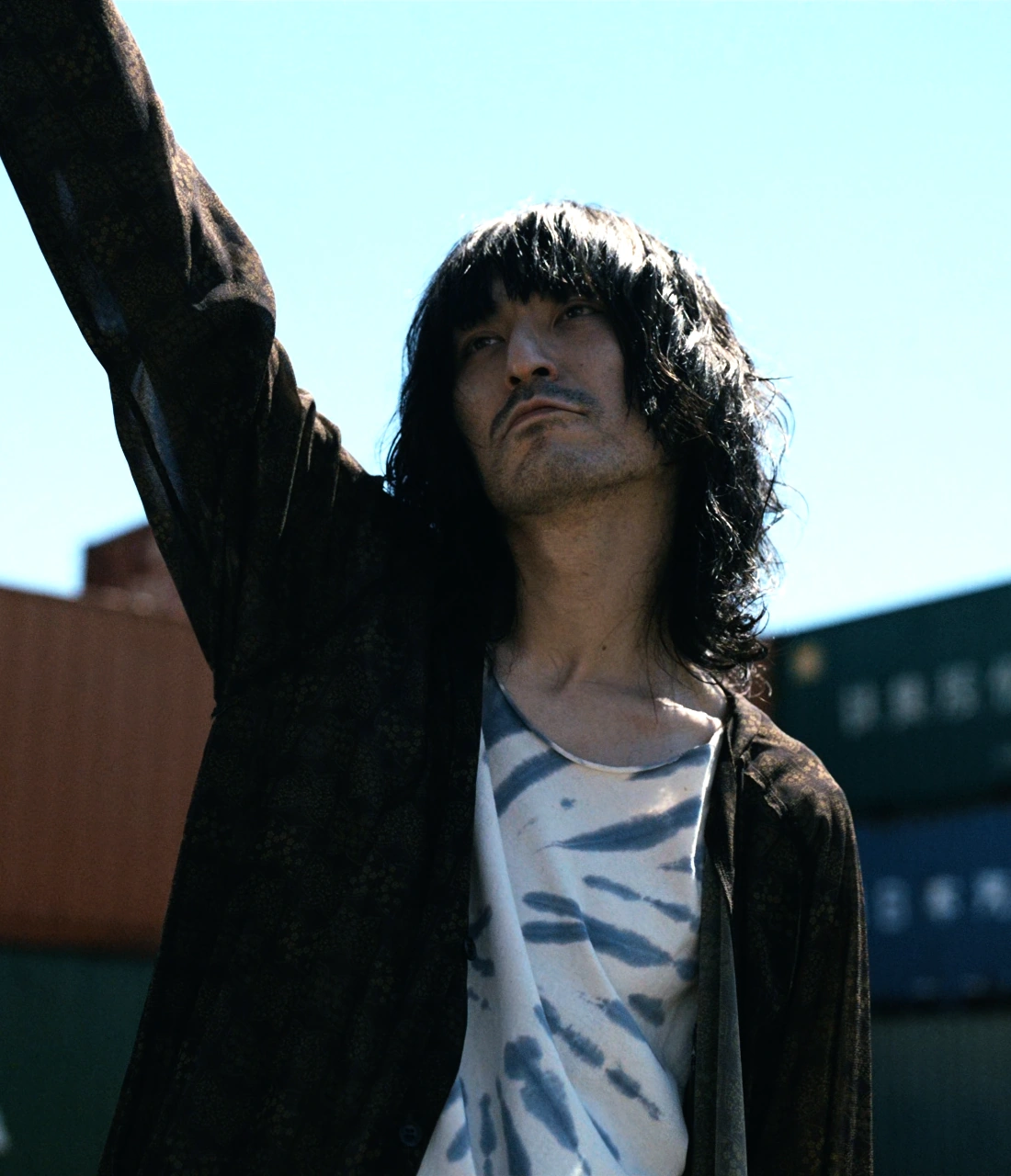 A screenshot of Shitara from Season 2, an older man with shoulder-length wavy hair, wearing a white shirt with a maroon cardigan on top