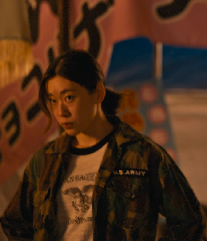 A woman with her hair in a ponytail, wearing a jacket with a military pattern and a white T-shirt underneath