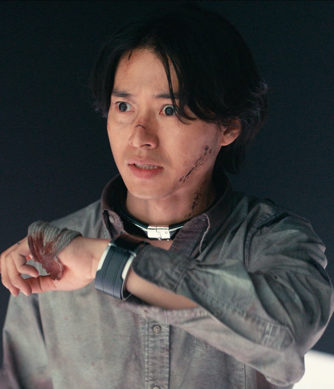 A close-up shot of Arisu staring forward in shock, his face bruised and hand bandaged, he is talking to someone through the wristband