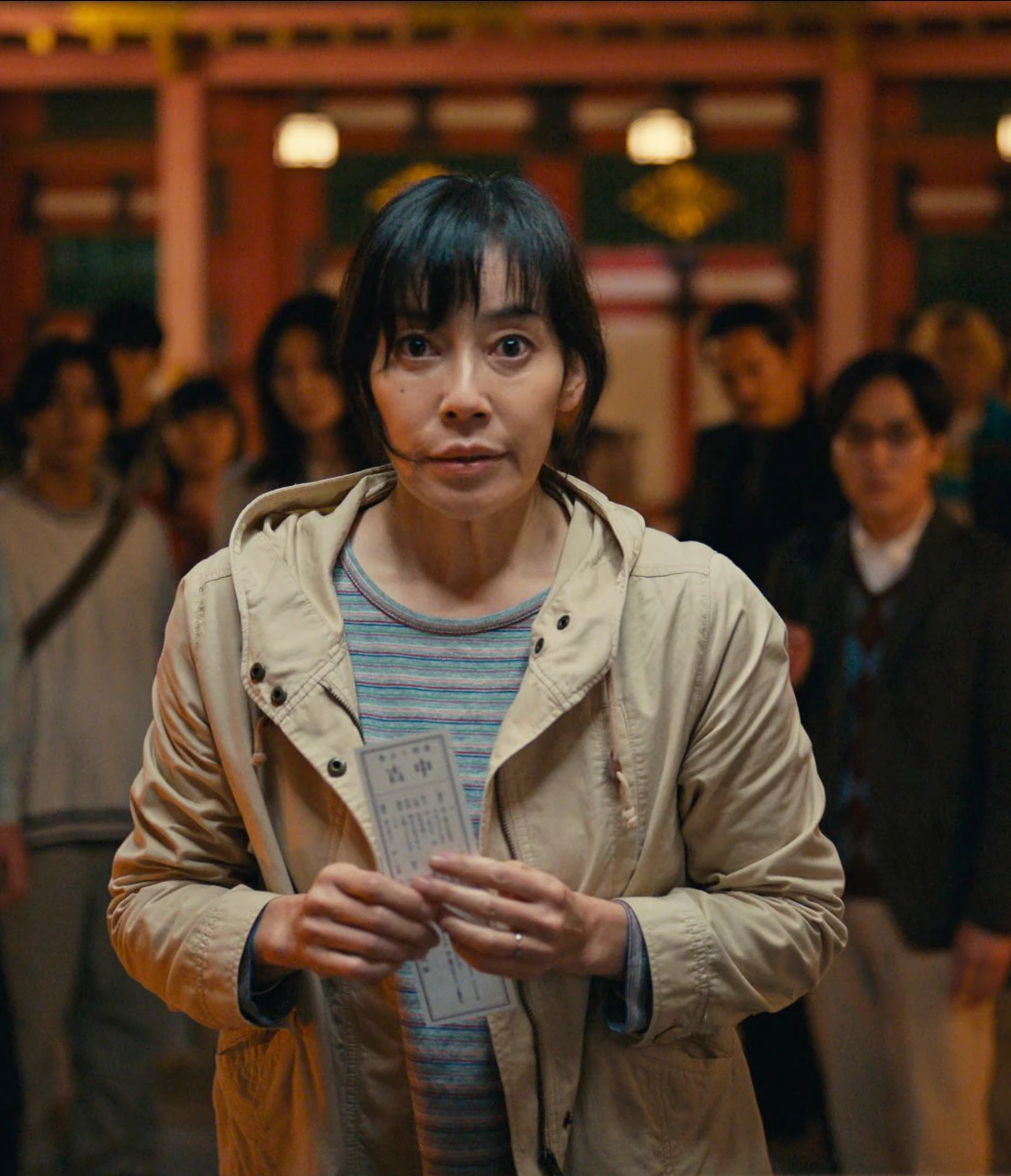 A close-up shot of Sachiko holding a fortune slip and looking at the game announcer with hope in her eyes, multiple other people are behind her