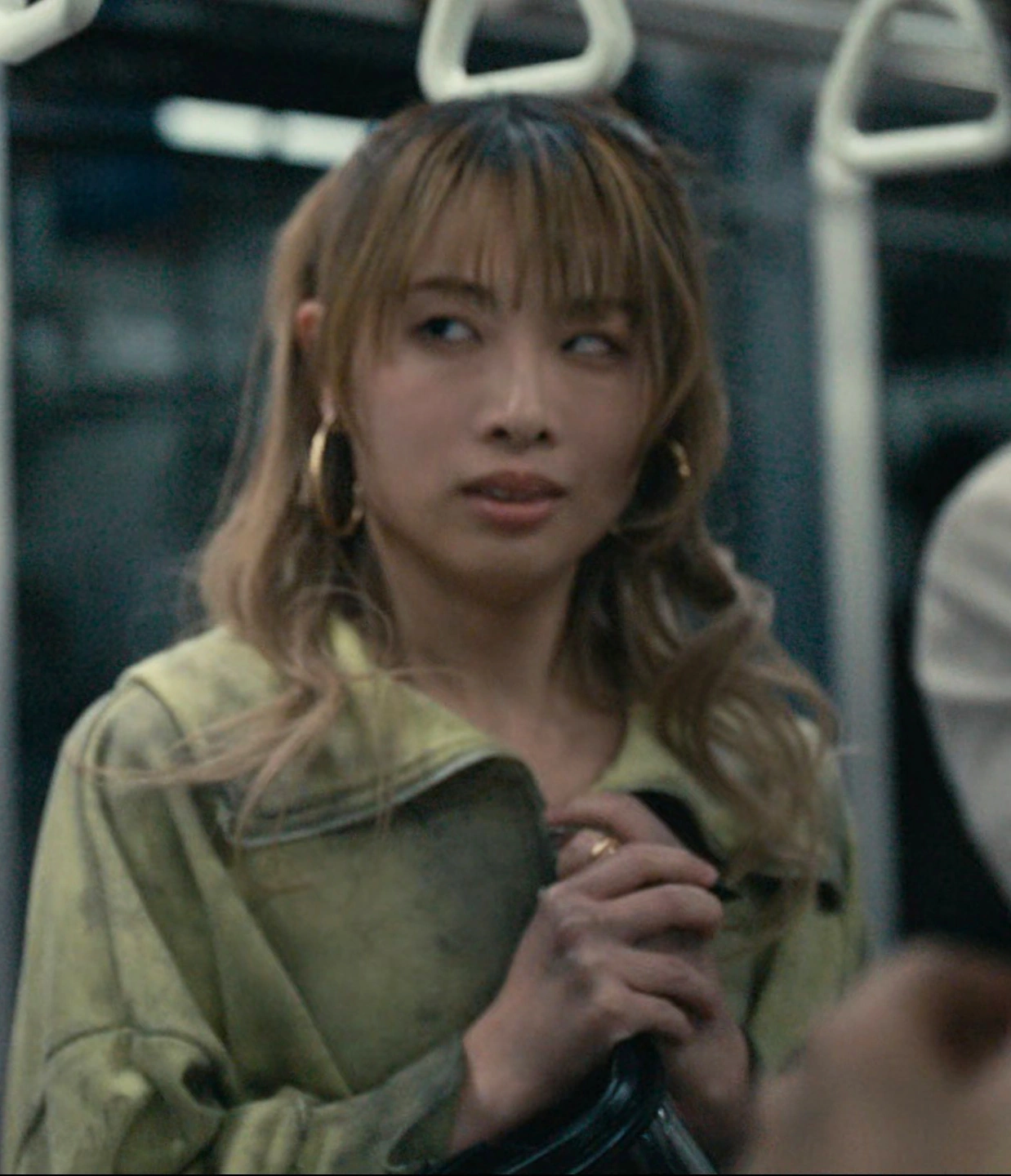 A screenshot of an unnamed female player with long blond hair and big round earrings, wearing a yellow/green jacket