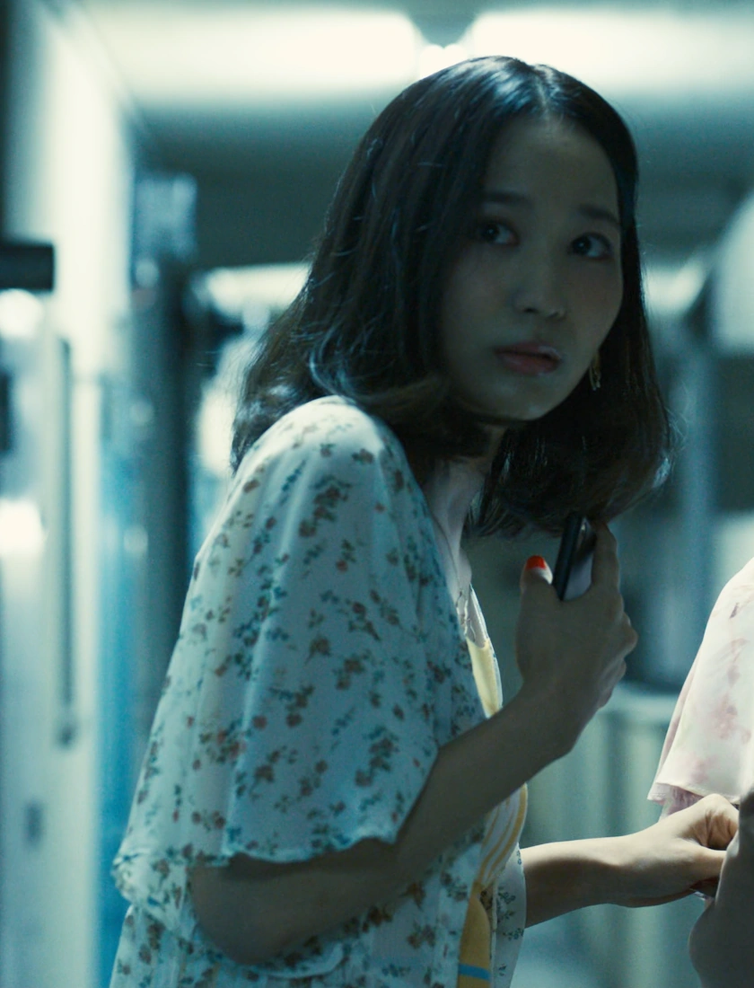 A screenshot of Aya from Season 1, a young woman with shoulder-length hair, wearing a yellow T-shirt with a white jacket on top