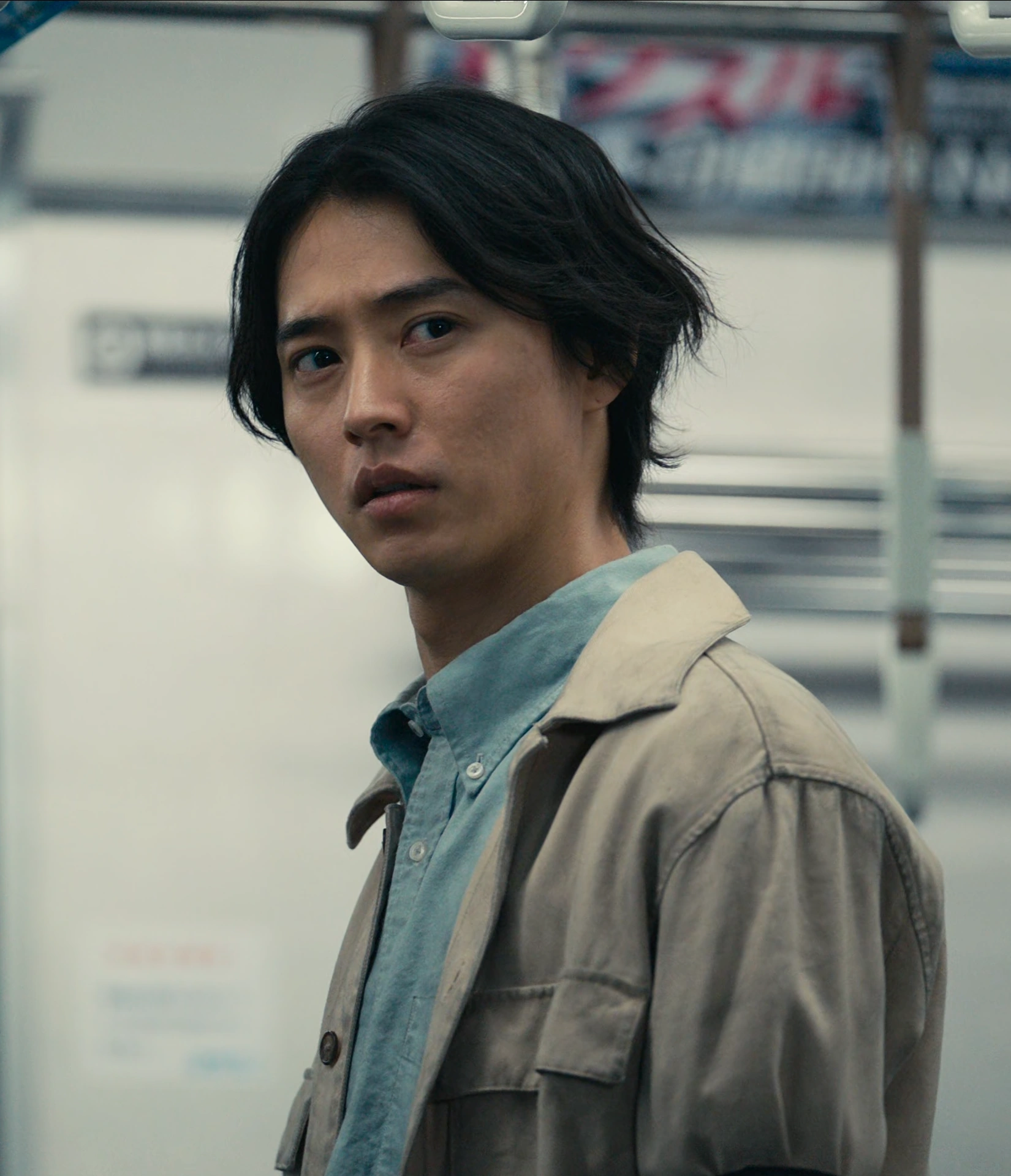 A screenshot of Ryohei Arisu from Season 3, a young man standing in the middle of a train in a beige coat, looking around suspiciously