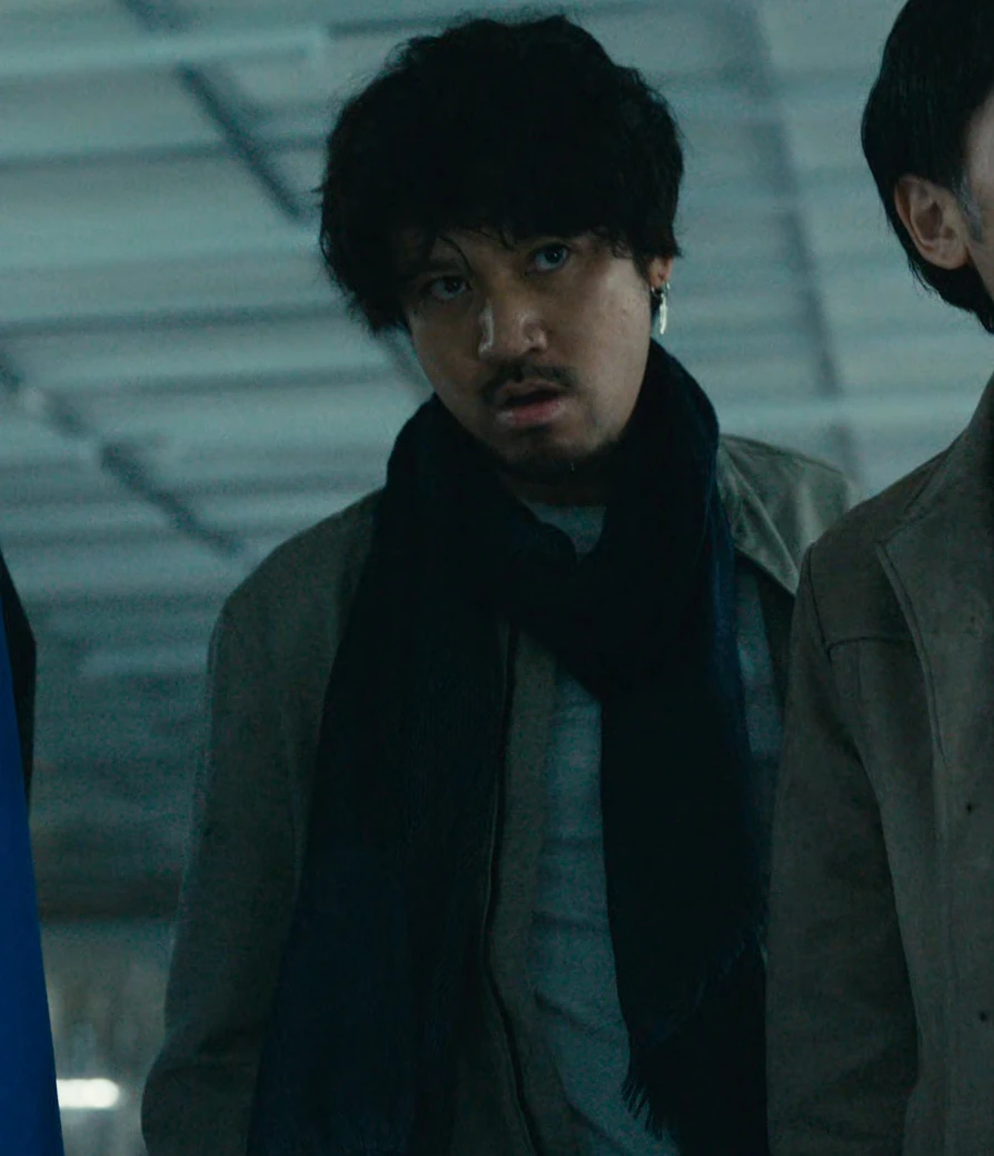 A middle-aged man with curly hair wearing a long scarf on top of a gray jacket with a blue shirt underneath