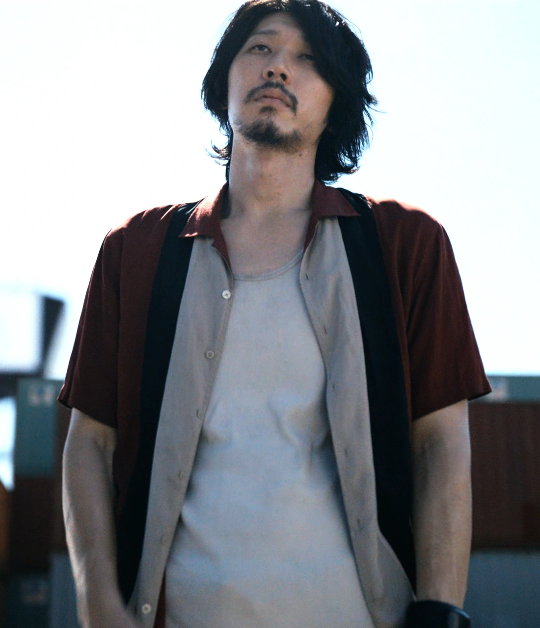 A screenshot of Maki from Season 2, a middle-aged man with a moustache wearing a red shirt over a white T-shirt, keeping his hands in his pockets