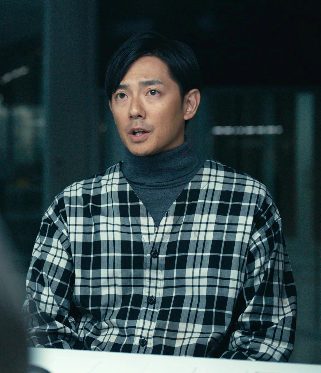 A middle-aged man with a medium-length black hair and bangs combed to the right side. He is dressed in a dark gray turtleneck sweater under a checked shirt