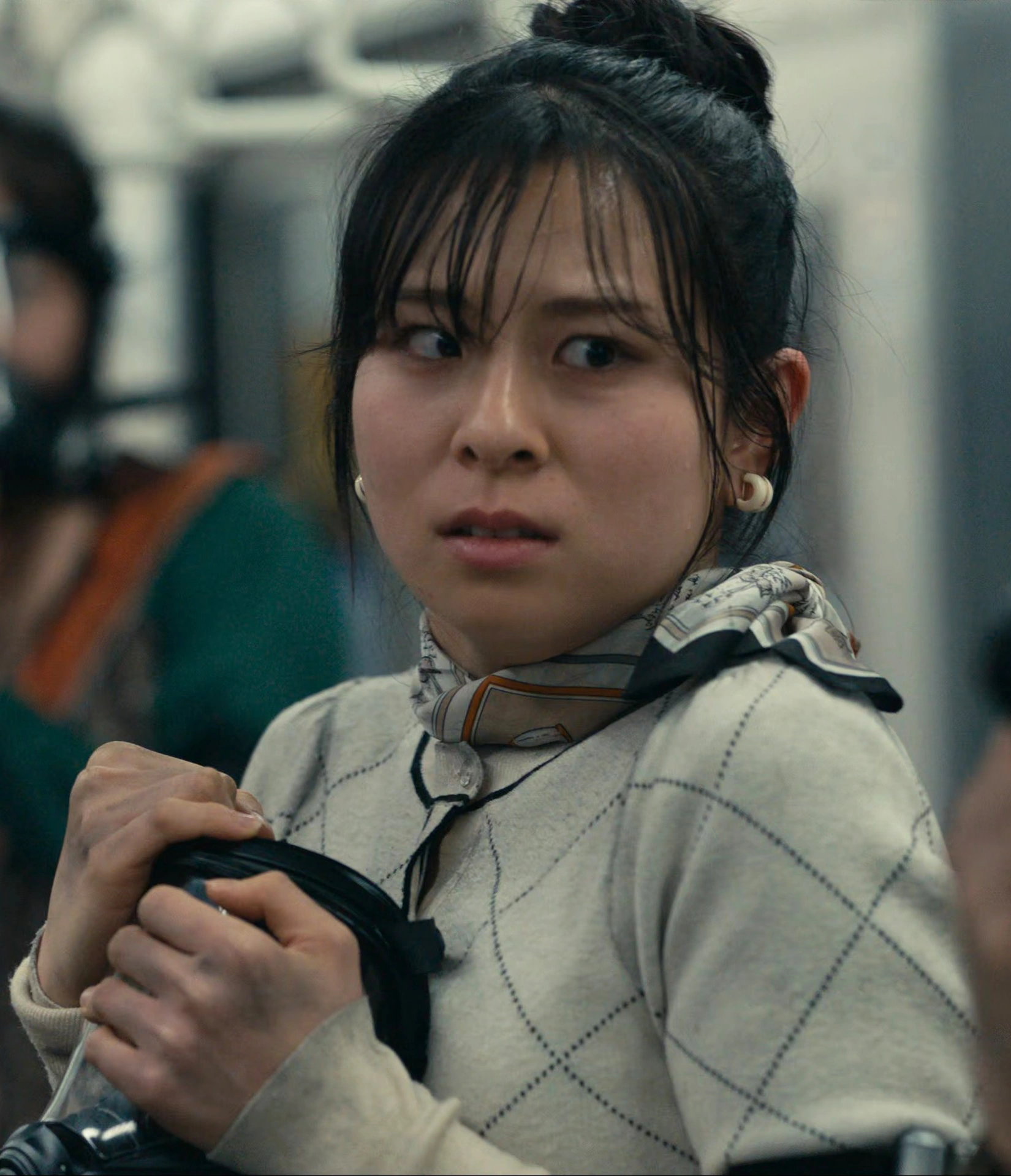 A screenshot of an unnamed female player with her hair in a bun, wearing a smart beige jacket and a scarf, looking terrified