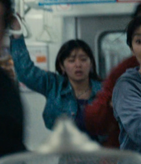 A screenshot of Natsu from Season 3, a young woman with medium-length hair wearing a denim jacket, holding onto a strap with a worried expression