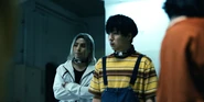Chishiya standing with his arms crossed and looking towards Ippei, while Furuta, dressed in orange, is blurred in the foreground