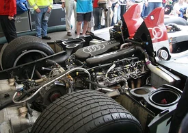 The Audi R10's exposed engine bay, viewed from the rear on a three-quarters angle. It shows the carbon-fibre intake plenums, which are emblazoned with the four-ring Audi logo and the letters "V12 TDI". Radiators are situated to the left and right of the engine, with air intakes for the rear wheels directly rearwards of them. The transmission and engine are both mounted longitudinally, with the former right behind the engine. The diesel particulate filters are immediately to the behind of the transmission.