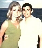 Sharon Tate and Jay Sebring photographed in 1966. They are standing side by side with an ocean background. Tate is wearing a sleeveless dress and Sebring a white shirt. They are standing close together. Sebring has his arm around Tate and they are looking directly into the camera, smiling.