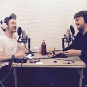 Griffin and David smiling and sitting across from one another, each wearing headphones and sitting in front of a microphone.