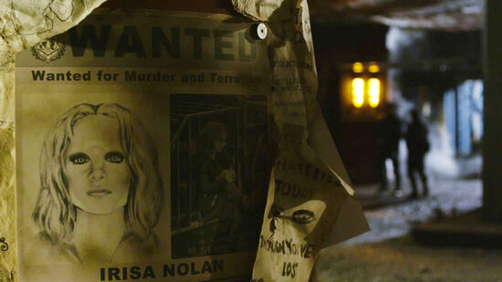 a wanted poster hung on a post stating "Wanted for Murder and Terrorism" above a drawing and photo of Irisa and her name "Irisa Nolan" written below her pictures