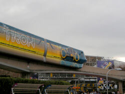 A monorail stamping a painted Light Cycle which leaves an orange trail behind. The second cart also has the Tron Legacy title.