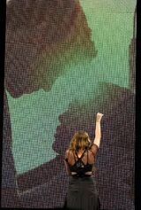 A female teenager with long brown hair and a black dress stretches her fist upward. Only the teen's back is visible, as she is facing a large screen behind her. On the screen is the silhouette of a male teen caressing the face of a shorter female teenager.Cyrus sings "When I Look at You" during her Wonder World Tour while facing a screen playing a film clip of Will and Ronnie.