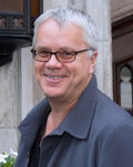 Tim Robbins—a white male with close-cut gray hair, light-colored eyes, smiling and wearing narrow, plastic-framed eyeglasses and a dark gray jacket over a black shirt—attending the Toronto International Film Festival in September 2012 at age 53.