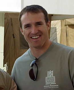 A man is smiling. He is wearing a gray T-shirt with a pair of black sunglasses hanging from the collar.
