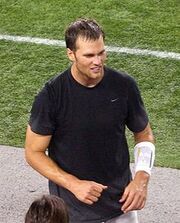 A man standing on the sidelines of an American football field. He is wearing a black shirt.